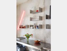 Moderne minimalistische Küche mit einer Arbeitsfläche aus Edelstahl, offenen Regalen aus Metall an einer beigen Wand und einem Dekoelement in Form eines leuchtenden, roten Neon-Lichts. Auf der Arbeitsfläche stehen eine Schale mit Obst, eine Vase mit Tulpen und ein tragbares Kochfeld.