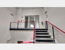 Treppenhaus in einem modernen Gebäude mit Betonstufen, rotem Handlauf und Glasgeländern. Im Hintergrund befindet sich eine Tür mit Glaseinsatz, durch den ein rundes Fenster sichtbar ist.