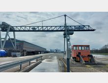 Industriehafen mit großen Metallkränen und einer langen Lagerhalle auf der linken Seite. Im Vordergrund steht eine orangefarbene Rangierlokomotive, während im Hintergrund weitere Hafeninfrastruktur und ein Fluss zu sehen sind.