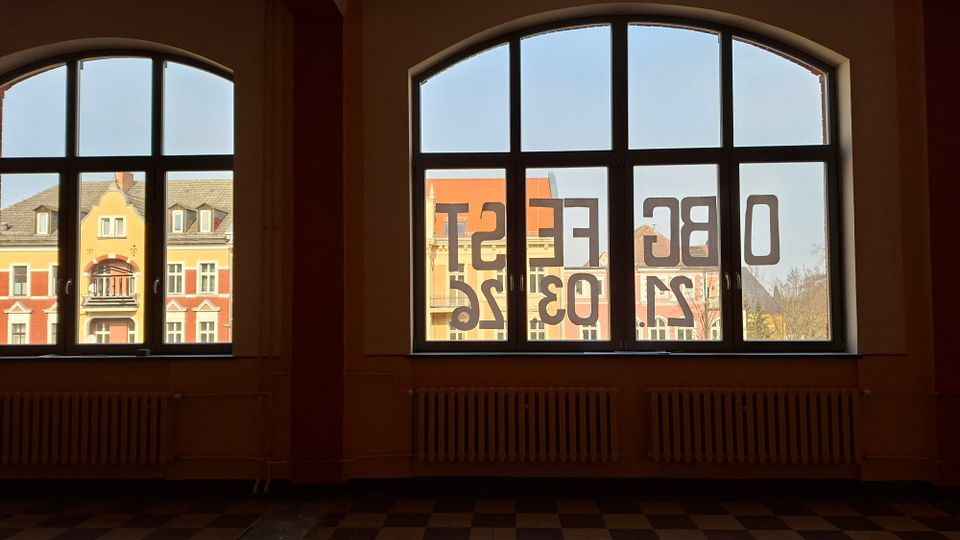 Blick aus einem Innenraum mit zwei großen, segmentbogigen Sprossenfenstern und Heizkörpern darunter. Durch die Fenster sieht man gegenüberliegende Wohnhäuser mit farbigen Fassaden und Satteldächern; auf dem rechten Fenster sind spiegelverkehrte Ziffern und Buchstaben aufgeklebt.