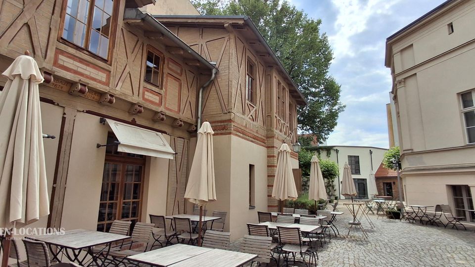 Gepflasterter Innenhof mit Café-Bestuhlung und mehreren geschlossenen Sonnenschirmen, flankiert von einem Fachwerkgebäude mit großen Fenstern und einem hellen Putzbau. Im Hintergrund stehen Bäume und weitere niedrige Gebäude, wodurch der Hof wie ein kleiner, geschützter Platz wirkt.