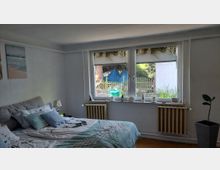 Schlafzimmer mit großem Doppelbett, hellen Wänden und Holzfußboden. Eine breite Fensterfront mit Fensterbankdekoration und zwei Heizkörpern blickt in einen kleinen Garten mit Zaun und Pflanzen.