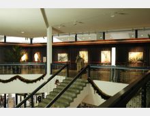 Innenraum eines modernen Gebäudes mit einer Treppe, dekoriert mit Weihnachtsgirlanden. Im Hintergrund befinden sich dunkle Vorhänge, mehrere abstrakte Gemälde und eine hohe Lampe, eingerahmt von Fenstern mit rechteckigen Lichtöffnungen.