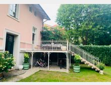 Helles Gebäude mit rosa Fassade und weißen Fensterrahmen, ergänzt durch einen erhöhten Balkon mit Metallgeländer und einer seitlich führenden Treppe. Der Gartenbereich darunter ist mit Fahrrädern, Pflanzkübeln und einem Steinweg gestaltet; im Hintergrund stehen Bäume und eine Hecke.