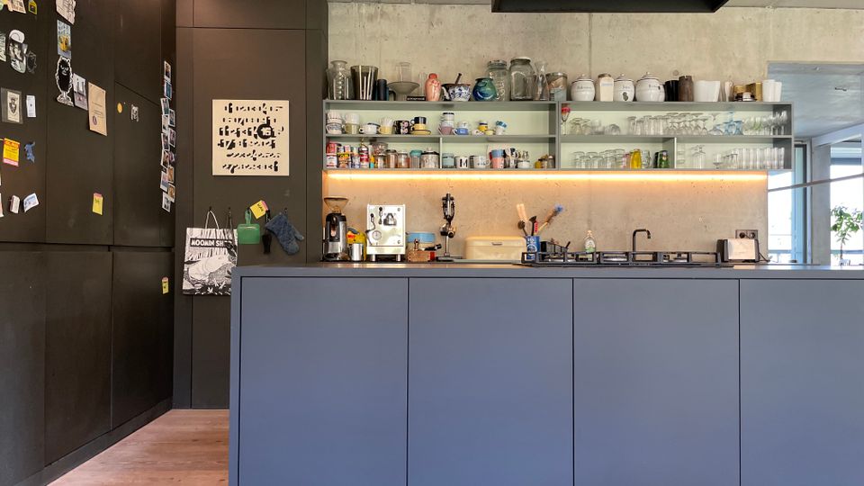 Moderne Küche mit einer Front aus matten, grauen Schränken, einer Betonwand mit offenen Regalböden, auf denen Geschirr und Gläser stehen, und einer Arbeitsplatte mit Küchengeräten und einem Kochfeld. Rechts hängt eine schwarze Dunstabzugshaube, links ist eine Wand mit dunklen Magneten und Dekorationen.