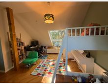 Ein Dachzimmer mit schrägen Wänden, einem Hochbett aus Holz, einem Schreibtisch mit Computer, einem Bücherregal, einem grünen Sessel und einer bunten Teppichmusterung. Ein großes Dachfenster lässt Tageslicht ein, während ein Klavier an der Wand steht.