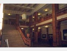 Ein historischer Innenraum mit einer zentralen Treppe, Holzgeländern und Balken in dunklem Holzton. Die Decke ist dekorativ gestaltet, und an den Wänden befinden sich kleine, eingebaute Lampen.