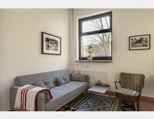 Wohnzimmer mit einer grauen Couch, einem kleinen Beistelltisch und einem grünen Sessel. Ein großes Fenster mit schwarzem Rahmen und einer Zimmerpflanze auf der Fensterbank dominiert den Raum, ergänzt durch schwarze gerahmte Fotografien an den Wänden.