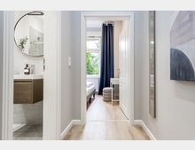 Heller, moderner Wohnungsflur mit hellen Holzdielen und weißen Türrahmen, Blick durch eine offene Tür in ein Schlafzimmer mit großem Fenster und dunkelblauem Vorhang. Links ist ein kleines Bad mit Waschtisch und rundem Spiegel zu sehen.