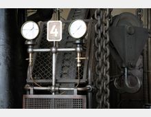 Industrielle Maschine mit zwei Zifferblättern zur Druckanzeige, einer großen Metallkette und einem massiven Haken. Im Hintergrund sind Gitter und weitere mechanische Elemente einer industriellen Anlage sichtbar.