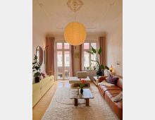 Wohnzimmer mit hellen Holzböden, hohen Decken und einer schlichten Stuckrosette, aus der eine große, runde Papierlampe hängt. Der Raum ist gemütlich eingerichtet mit einem braunen Ledersofa, einem zentralen Marmortisch, Pflanzen und großen Fenstern mit Vorhängen, die zu einem Balkon führen.