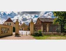 Historische Hofanlage mit gelbem Eingangstor und Kopfsteinpflaster, umgeben von Gebäuden mit Ziegeldächern; im Hintergrund ist ein Kirchturm zu sehen. Rechts steht ein Zaun, der eine Statue und Grünflächen umgibt.