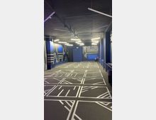 Langer, moderner Innenraum mit dunkelblauen Wänden und grauem Boden, der mit weißen geometrischen Linienmustern gestaltet ist. An der schwarzen Decke hängen mehrere lineare LED-Leuchten; am Ende des Raums befindet sich eine blaue Tür.