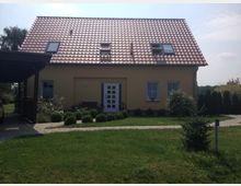 Einfamilienhaus mit gelber Fassade und rotem Satteldach, zwei Dachfenstern und einem zentralen Eingang mit Glastür. Vor dem Haus ein gepflegter Garten mit Gehweg, Buchsbaumhecken und einzelnen Bäumen; links ein überdachter Carport.