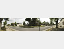 Straßenkreuzung in einer Vorortsiedlung mit älteren Wohnhäusern, teils aus Ziegel- und Putzfassaden, umgeben von Bäumen und Grünflächen. Der Gehweg aus Pflastersteinen verläuft entlang der Straße, und einzelne parkende Fahrzeuge sind im Hintergrund zu sehen.