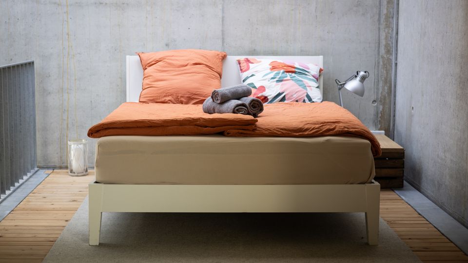 Modernes Schlafzimmer mit einer minimalistisch gestalteten weißen Bettgestell auf einem Holzfußboden. Das Bett ist mit orangefarbener Bettwäsche, einem Kissen mit floralem Muster und gerollten Handtüchern dekoriert; die Betonwand im Hintergrund verleiht dem Raum einen industriellen Look.