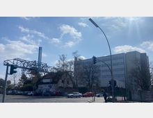 Straßenszene mit einem mehrstöckigen, weißen Bürogebäude im Hintergrund, einem kleineren, verfallenen Gebäude mit Graffiti im Vordergrund und einer großen Stahlkonstruktion links. Eine Ampel und parkende Autos sind ebenfalls sichtbar, umgeben von Bäumen unter einem leicht bewölkten Himmel.