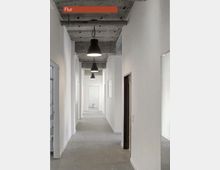 Langer, schlichter Flur mit weißen Wänden, grauem Betonboden und einer offenen Decke aus Sichtbeton. An der Decke hängen mehrere schwarze Pendelleuchten in regelmäßigen Abständen.