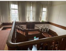 Treppenhaus eines historischen Gebäudes mit Holzgeländern, einem großen Kronleuchter und hohen Fenstern mit weißen Vorhängen. Die Wände sind weiß, mit holozontalen Holzverkleidungen, und das Treppenhaus führt in ein Erdgeschoss mit Schachbrettmuster-Fliesen.