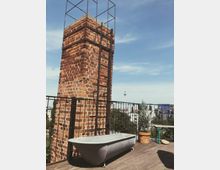 Rooftop-Terrasse mit Holzboden, einem freistehenden, alten Ziegelkamin mit Metallleiter und einer metallenen Badewanne im Vordergrund. Im Hintergrund sind Bäume, städtische Gebäude und der Berliner Fernsehturm sichtbar.