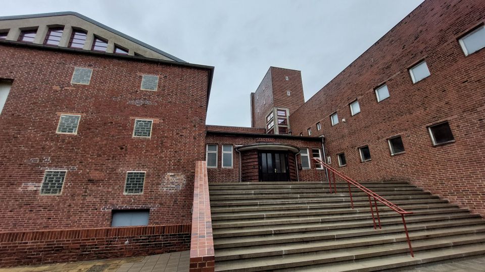 Backsteinbau mit modernistischem Design, bestehend aus mehreren Gebäudeteilen mit schlichten Fenstern. Eine breite Treppe mit rotem Geländer führt zum Eingangsbereich mit abgerundeter Überdachung, im Hintergrund ragt ein Turm empor.