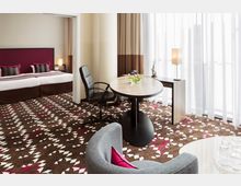 Ein modernes Hotelzimmer mit einem separaten Schlafbereich, das über ein großes Bett mit farblich abgestimmten Kissen, einen Schreibtisch mit Bürostuhl und ein rundes Beistelltischchen verfügt. Der Raum ist hell durch bodenlange Vorhänge, die Tageslicht einlassen, und der gemusterte Teppich setzt farbliche Akzente.