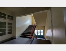 Treppenhaus eines Gebäudes mit beige gestrichenen Wänden, dunkelbraunen Stufen und einem roten Geländer. Im Hintergrund befindet sich eine Tür mit mehreren Glasfeldern, die zu einem Ausgang führt.