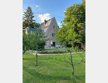 Ein Einfamilienhaus mit spitzem Dach und Solarpaneelen, umgeben von einem gepflegten Garten mit grüner Rasenfläche, kleinen Bäumen und einer Terrasse mit Sitzmöbeln und Sonnenschirm. Im Hintergrund befinden sich hohe Bäume unter einem sonnigen Himmel.