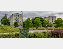 Mehrstöckiges Wohnhaus mit einer auffälligen Wandbemalung, die Figuren und Alltagsgegenstände zeigt, umgeben von Bäumen und Grünflächen. Im Vordergrund befindet sich ein Kreisverkehr mit Verkehrsschildern, darunter ein Wegweiser nach Lübben.