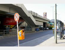 Straßenszene mit einer Bushaltestelle im Vordergrund, einem orangefarbenen Mülleimer und einem "Halteverbot"-Schild. Im Hintergrund sind eine erhöhte Straßenbrücke, ein Friseursalon mit roter Fassade und ein Backsteingebäude mit Berliner Wappen zu sehen.