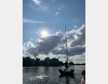 Segelboot auf einem ruhigen Gewässer, mit einem stehenden Mann an Bord, umgeben von bewaldetem Ufer. Der Himmel ist teils wolkig, und die Sonne scheint durch die Wolken hindurch.