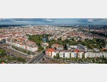 Luftaufnahme einer städtischen Landschaft mit dichter Wohnbebauung, Parks und einem kleinen See in der Mitte. Im Vordergrund sind Bahngleise und Straßenkreuzungen zu sehen, während am Horizont moderne Hochhäuser hervorragen.