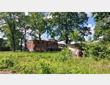 Ziegelsteingebäude in Ruinen mit eingeschlagenen Fenstern, umgeben von dichter Vegetation und Bäumen; im Vordergrund ist eine überwucherte Wiese zu sehen. Teile der Gebäudefassade sind mit Graffiti bemalt.