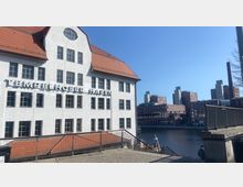 Weißes Gebäude mit der Aufschrift "Tempelhofer Hafen", roten Dachziegeln und zahlreichen Fenstern, im Hintergrund mehrere moderne Backsteingebäude an einem Wasserkanal unter klarem, blauem Himmel. Im Vordergrund führt eine Metalltreppe entlang einer gepflasterten Fläche.