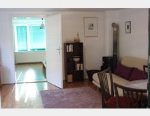 Wohnzimmer mit Holzboden und weißen Wänden, ausgestattet mit einem hellen Sofa mit violetten Kissen, einem kleinen runden Esstisch und Stühlen. Im Hintergrund ist eine geöffnete Tür zu einem weiteren Raum mit Fenster und grünen Jalousien sichtbar.