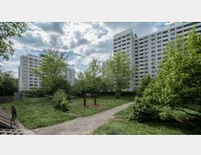 Weitläufige Grünfläche mit Rasen, Bäumen und einem kleinen Spielplatz mit einer Schaukel im Vordergrund, umgeben von mehreren hohen Wohnblöcken aus Beton sowie bewölktem Himmel im Hintergrund.