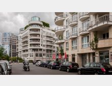 Straßenszene mit modernen Wohn- und Bürogebäuden, die durch helle Fassaden mit großen Fenstern und geschwungenen Balkongeländern gekennzeichnet sind. Geparkte Autos und ein Motorroller befinden sich am Straßenrand, während im Hintergrund ein rundes, verglastes Hochhaus sichtbar ist.