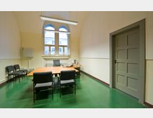 Ein einfacher Besprechungsraum mit grünem Boden, heller Wandgestaltung und hohen, bogenförmigen Fenstern. Im Raum stehen ein Holztisch, mehrere schwarze Stühle sowie ein Schreibtisch mit Bürotechnik; eine graue Tür befindet sich an der rechten Seite.