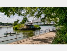 Eine Flusslandschaft mit einer Brücke im Vordergrund, die von weißen Stahlbögen getragen wird. Unter der Brücke paddeln drei Personen in einem Kanu, während am Flussufer ein Gehweg mit Geländer verläuft und von Bäumen gesäumt wird.