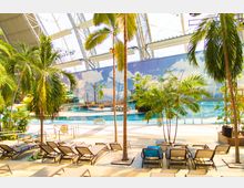 Innenansicht eines tropischen Freizeitparks mit einem Pool, umgeben von Palmen und Liegestühlen. Im Hintergrund sind eine aufgemalte Himmelkulisse, Wasserrutschen und Holzstege sichtbar, unter einem weit gespannten Dach mit Stahlträgern.