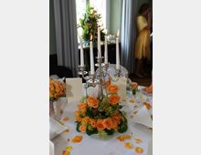 Ein festlich gedeckter Tisch mit einem silbernen Kerzenständer mit brennenden weißen Kerzen, umgeben von einem Blumenarrangement aus orangefarbenen Rosen und grünem Laub. Im Hintergrund sind Vorhänge, eine Vase mit Blumen und eine Person in gelbem Kleid zu sehen.