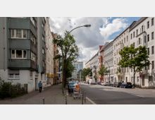 Straßenszene in einer Wohngegend mit mehrstöckigen Altbau- und modernen Gebäuden auf beiden Seiten. Entlang der Straße stehen geparkte Autos, vereinzelte Bäume und ein Gehweg mit Kopfsteinpflaster; im Hintergrund ein modernes Glasgebäude und blauer Himmel mit Wolken.