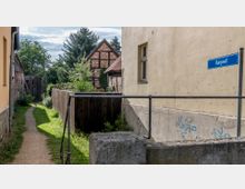 Schmaler Weg entlang eines Zauns und einer gelben Hausfassade mit dem Straßenschild „Burgwall“; im Hintergrund ein Fachwerkhaus, umgeben von Bäumen und Gärten.