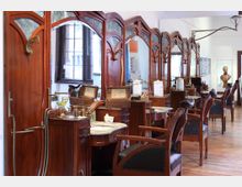 Raum mit einer Reihe historischer Friseurstühle und -spiegel aus Holz mit geschwungenen Verzierungen, ergänzt durch Marmorwaschbecken und antike Messingarmaturen; im Hintergrund ein großes Fenster und eine Büste auf einem Sockel.