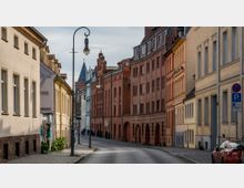 Straßenszene in einer historischen Altstadt mit Gebäuden aus Ziegel- und Putzfassaden in warmen Farbtönen; im Hintergrund ragen Kirchtürme hervor, und Laternen im klassischen Stil säumen den Gehweg.