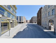 Straßenszene mit moderner und historischer Architektur, bestehend aus Gebäuden mit großen Glasfassaden, klassischen Fassadenelementen und einem weiten, asphaltierten Platz. Im Hintergrund sind weitere mehrstöckige Gebäude in symmetrischer Anordnung sichtbar.