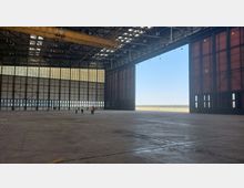 Große, leere Flugzeughalle mit hohen Decken, Stahlträgern und einem überdimensionalen geöffneten Tor, das den Blick auf eine Landebahn und den Himmel freigibt. Im Vordergrund sind mehrere Personen in Warnwesten zu sehen, die in der Halle stehen.