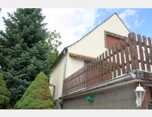 Zweistöckiges Wohnhaus mit cremefarbener Fassade, braunen Fenstern mit Rollläden und einem Holzzaun auf der erhöhten Terrasse. Im Vordergrund befinden sich Nadelbäume, während eine weiße Außenlaterne am rechten unteren Rand sichtbar ist.
