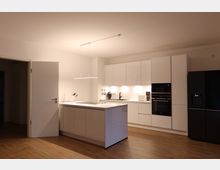 Moderne Küche mit weißen, glänzenden Schränken und integrierten Elektrogeräten, einer zentralen Kücheninsel und Holzfußboden. Links eine geöffnete Tür, rechts ein schwarzer Kühlschrank, die Umgebung ist minimalistisch gestaltet.