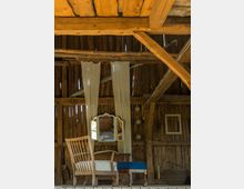 Holzinterieur eines rustikalen Raumes mit sichtbaren Holzbalken und Bretterwänden. Im Vordergrund steht ein Holzstuhl neben einer Sitzbank, dahinter ein Holzspiegel auf einem kleinen Tisch, umrahmt von weißen Stoffbahnen.